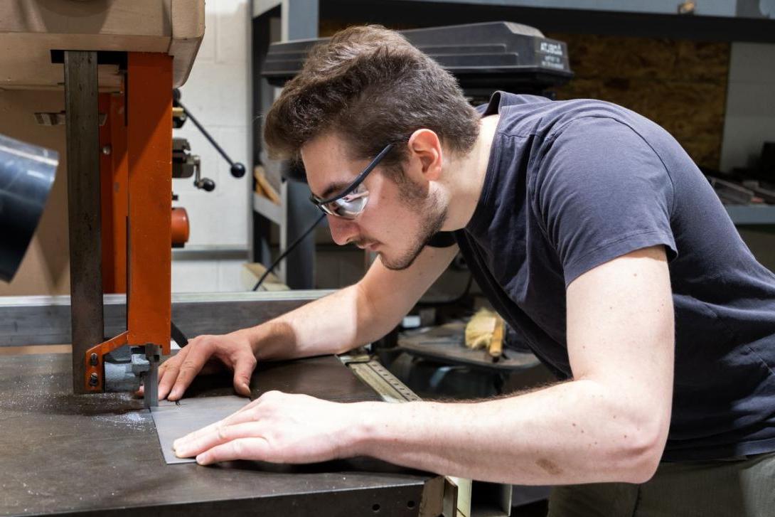 A Kettering student wearing safety glasses cuts a piece of metal with heavy equipment in a lab space